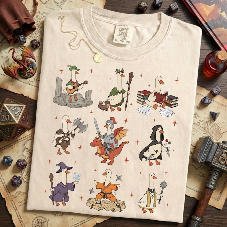 May include: Cream-colored t-shirt featuring cartoon geese in various fantasy roles, such as a bard, a wizard, and a knight riding a dragon. The shirt has the text "Comfort Colors XL 70-20" printed on the upper right side. The design includes red star accents.