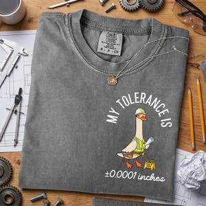 May include: A gray Comfort Colors t-shirt with the text "MY TOLERANCE IS ±0.0001 inches" and a cartoon goose wearing a hard hat and safety vest. The shirt has a crew neck and short sleeves.