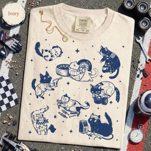 May include: Ivory-colored t-shirt featuring navy blue cartoon cats in racing attire, with tools and tires. The shirt has a crew neck and the Comfort Colors XL 78-20 label. A gold necklace and racing accessories are on the surface.