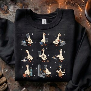 May include: Black sweatshirt featuring a repeating pattern of cartoon geese in welding gear. Each goose is depicted in a different welding pose, with sparks and stars around them. The geese are wearing welding helmets and holding tools.