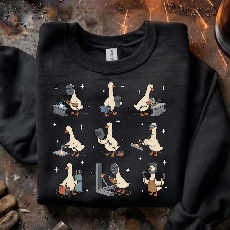 May include: Black sweatshirt featuring a repeating pattern of cartoon geese in welding gear. Each goose is depicted in a different welding pose, with sparks and stars around them. The geese are wearing welding helmets and holding tools.