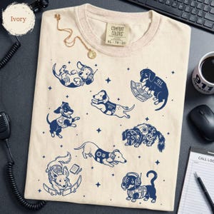 May include: An ivory t-shirt featuring a navy blue graphic print of various dogs wearing headsets and 911 dispatcher gear. The shirt has a Comfort Colors tag at the neck, size XL, and is decorated with small star icons scattered among the dog illustrations.