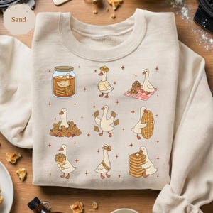 May include: Sand-colored sweatshirt featuring a whimsical design of cartoon geese interacting with waffles. The geese are depicted in various scenes, including enjoying waffles, carrying them, and in a jar. The design is playful and charming.