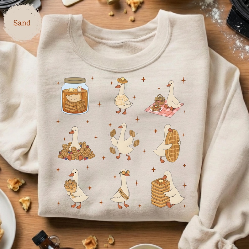 May include: Sand-colored sweatshirt featuring a whimsical design of cartoon geese interacting with waffles. The geese are depicted in various scenes, including enjoying waffles, carrying them, and in a jar. The design is playful and charming.
