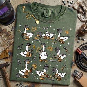 Pode incluir: Camiseta verde com um padr&atilde;o repetido de gansos de desenho animado soldando. Cada ganso &eacute; retratado com uma m&aacute;scara de solda, luvas e macac&atilde;o, segurando um ma&ccedil;arico de solda. A camisa tem um visual desgastado e gola redonda.