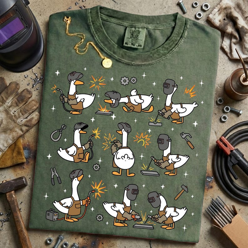 Pode incluir: Camiseta verde com um padr&atilde;o repetido de gansos de desenho animado soldando. Cada ganso &eacute; retratado com uma m&aacute;scara de solda, luvas e macac&atilde;o, segurando um ma&ccedil;arico de solda. A camisa tem um visual desgastado e gola redonda.