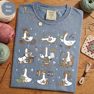 May include: Blue jean Comfort Colors t-shirt featuring a whimsical illustration of geese engaged in various crafts like knitting, crocheting, and spinning yarn. The shirt has a crew neck and a tag that reads XL 78-20. Yarn and crochet hooks are arranged around the shirt.