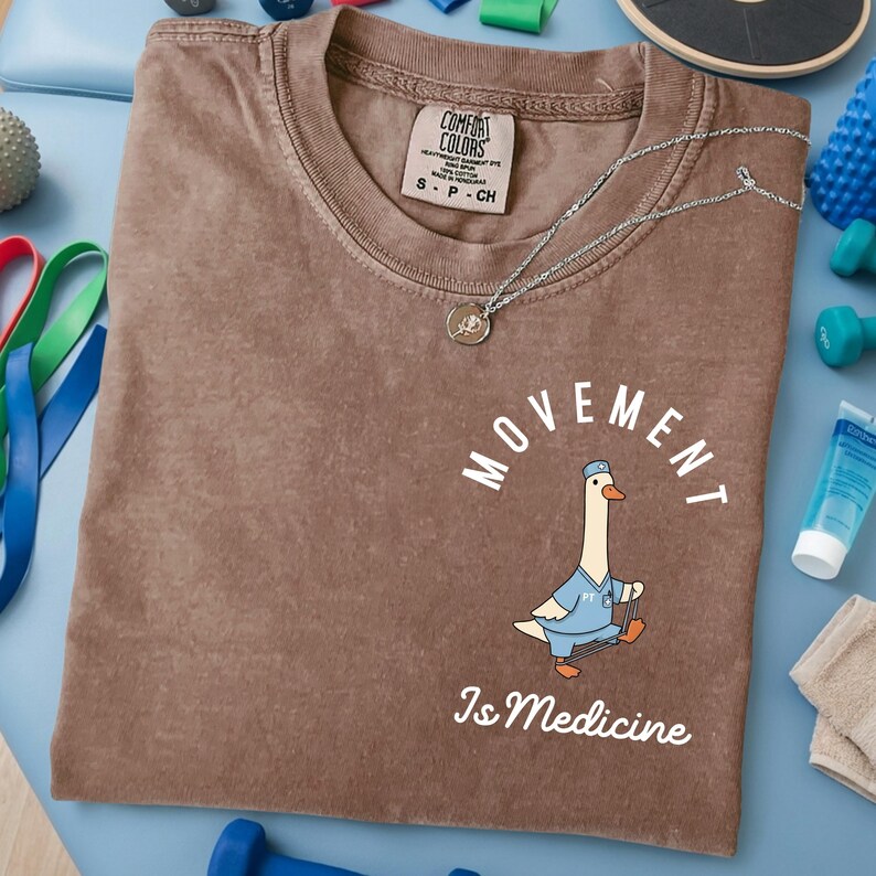 May include: Brown Comfort Colors t-shirt with the text "MOVEMENT Is Medicine" and a cartoon goose wearing a doctor's uniform. A silver necklace with a pendant rests on the shirt. The shirt is surrounded by exercise equipment.