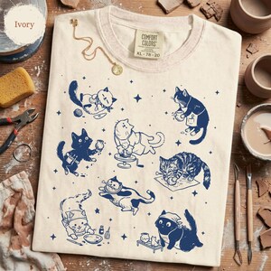 May include: Ivory-colored t-shirt featuring a playful design of cats engaged in pottery. The cats are depicted in navy blue, with various poses and tools. The shirt includes the text "COMFORT COLORS" and size "XL 78-20".