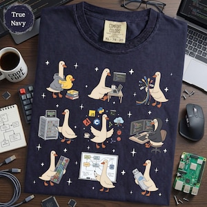 May include: A navy blue t-shirt featuring a graphic print of white geese engaged in various computing tasks, including coding, managing server racks, and handling cables. The shirt has a Comfort Colors label at the neck and a True Navy color tag.