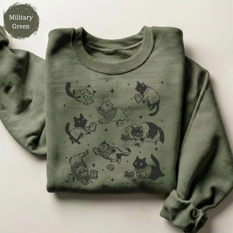 May include: A military green long-sleeve sweatshirt featuring a black graphic print of multiple cats in various poses, some wearing glasses or clothing, surrounded by small star shapes and books, with a crew neckline and ribbed cuffs.