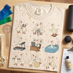 Pode incluir: Camiseta creme Comfort Colors XL 70-20 com gansos de desenho animado em v&aacute;rios cen&aacute;rios de fisioterapia. A camisa inclui gansos usando uma esteira, halteres e uma prancha de equil&iacute;brio. Um colar de ouro est&aacute; drapeado na camisa.
