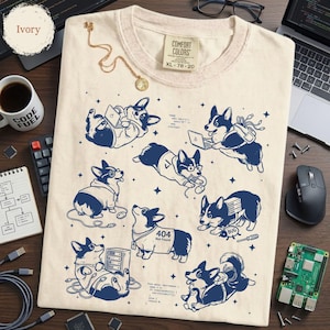 May include: An ivory t-shirt featuring a dark blue graphic print of multiple corgi dogs interacting with computer equipment and code. The design includes various corgis with laptops, cables, and a dog wearing a shirt labeled 404 Not Found.