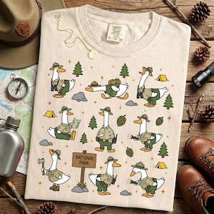 Op de afbeelding: Cr&egrave;mekleurig T-shirt met een grillig ontwerp van cartoonganzen verkleed als parkwachters. De ganzen worden afgebeeld in verschillende buitenactiviteiten, omringd door bomen, tenten en een "National Park"-bord. Het shirt heeft een label met XL-78-20.