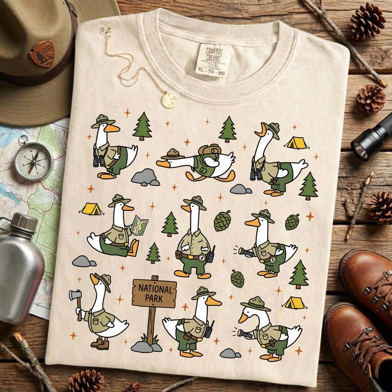Op de afbeelding: Cr&egrave;mekleurig T-shirt met een grillig ontwerp van cartoonganzen verkleed als parkwachters. De ganzen worden afgebeeld in verschillende buitenactiviteiten, omringd door bomen, tenten en een "National Park"-bord. Het shirt heeft een label met XL-78-20.