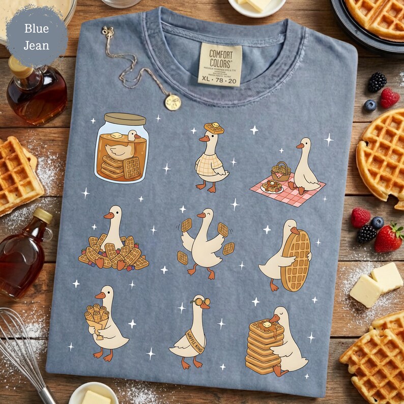 May include: Blue jean Comfort Colors t-shirt featuring a whimsical design of cartoon geese interacting with waffles and syrup. The geese are depicted in various poses, some wearing hats and glasses. The shirt includes the text "Waffle King".