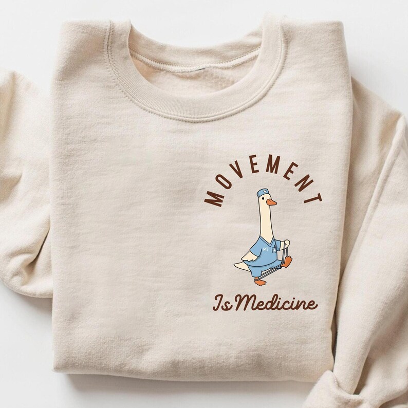 May include: A cream-colored sweatshirt featuring a cartoon goose in a blue medical uniform, holding a cane. The text "MOVEMENT" arches above the goose, with "Is Medicine" below in brown lettering. The sweatshirt is folded.