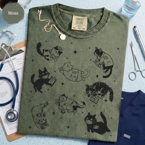 May include: Moss green t-shirt with a crew neck and short sleeves. The shirt features black illustrations of cats in medical roles, including a nurse, doctor, and wearing face masks. The tag reads "COMFORT COLORS XL 78-20".
