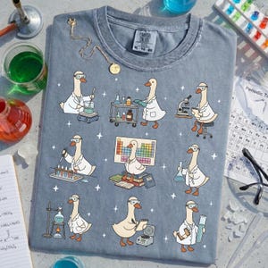 May include: A blue-grey t-shirt featuring cartoon geese in lab coats and safety goggles, engaged in various scientific activities. The design includes beakers, microscopes, and a periodic table. The shirt has a Comfort Colors tag.