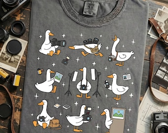 May include: Gray t-shirt featuring cartoon geese with cameras, flashes, and photo equipment. The geese are in various poses, some holding cameras, others with bags, and one with a press badge. The shirt has a crew neck and a vintage look.