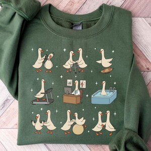Pode incluir: Camisola verde com um design caprichoso com gansos de desenho animado em v&aacute;rias atividades, incluindo boliche, usando um andador e fazendo exerc&iacute;cios. Os gansos s&atilde;o retratados em uma esteira, em uma mesa e em uma piscina. Estrelas brancas espalhadas.