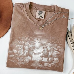 May include: Brown Comfort Colors t-shirt featuring a graphic of dancing frogs. The shirt has a crew neck and short sleeves. A silver necklace with a pendant rests on the shirt. The shirt is size small.
