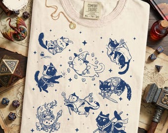 Könnte beinhalten: Elfenbeinfarbenes T-Shirt mit einem Design aus marineblauen Cartoon-Katzen in Fantasy-Rollen. Die Katzen werden als Zauberer, Bogenschützen und andere Charaktere dargestellt. Das Shirt hat einen Rundhalsausschnitt und kurze Ärmel. Das Etikett lautet "COMFORT COLORS XL 78-20".