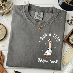 May include: Gray Comfort Colors t-shirt with a graphic of a goose in a captain's uniform and the text "I RUN A TIGHT Shipwreck". A silver necklace with a pendant is draped across the shirt.