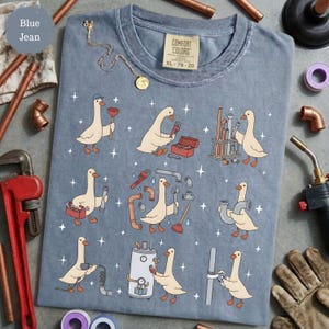 May include: A blue jean colored t-shirt featuring a graphic print of white geese performing various plumbing tasks with tools and pipes. The shirt has a Comfort Colors label at the neck with the text XL 78-20. The design includes small white star accents scattered among the geese.