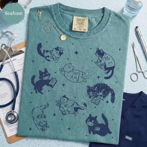 May include: Seafoam green t-shirt with a crew neck and short sleeves. The shirt features a pattern of navy blue cartoon cats in various medical roles, including nurses and doctors. The tag reads XL 78-20.