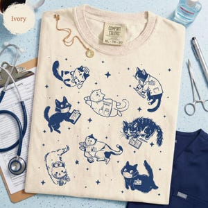 May include: Ivory-colored t-shirt with navy blue illustrations of cats in medical attire, including masks and lab coats. The shirt has the text "COMFORT COLORS" and size XL 78-20. A gold necklace and medical tools are also visible.