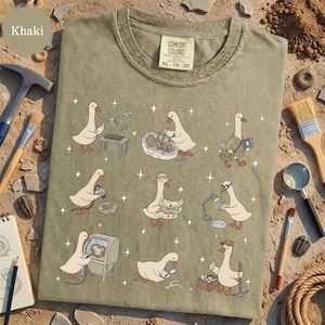 May include: Khaki-colored t-shirt featuring a whimsical design of cartoon geese engaged in paleontological activities. The geese are depicted excavating fossils, studying maps, and examining artifacts. The shirt has a Comfort Colors tag, size XL, and the number 78-20.