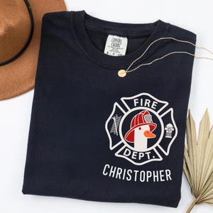 May include: Navy blue t-shirt with a white and red firefighter logo featuring a goose wearing a helmet. The text "FIRE DEPT." and "CHRISTOPHER" are printed below the logo. A gold necklace is draped across the shirt.