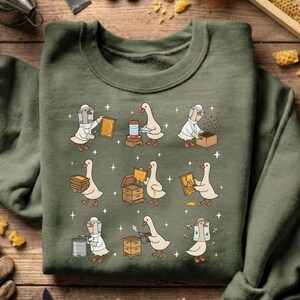 Puede incluir: Sudadera verde oliva con cuello redondo con un dise&ntilde;o caprichoso de gansos vestidos de apicultores. Los gansos se muestran trabajando con panales, tarros y colmenas. El gr&aacute;fico est&aacute; detallado con peque&ntilde;as estrellas.