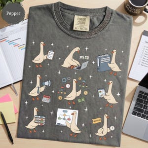May include: A charcoal gray t-shirt featuring a repeating pattern of cartoon geese in various office settings. The geese are depicted using laptops, holding clipboards, presenting charts, and managing files, interspersed with small white stars.