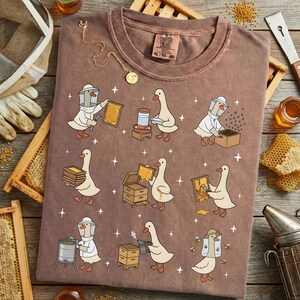 Puede incluir: Camiseta marr&oacute;n con un dise&ntilde;o caprichoso de gansos vestidos de apicultores. Los gansos se muestran trabajando con panales, colmenas y otros equipos de apicultura. La camiseta tiene cuello redondo y mangas cortas.