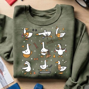 May include: Olive green sweatshirt featuring a repeating pattern of cartoon geese engaged in various exercises. The geese are depicted with weights, on a treadmill, and using resistance bands. The design includes orange, yellow, and black accents.