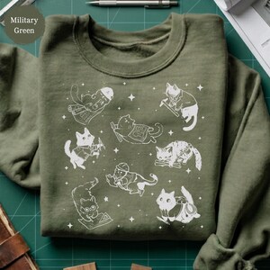 May include: Military green sweatshirt featuring a playful design of white cartoon cats in various occupations, such as a construction worker, a carpenter, and a pilot. The design includes stars and is printed on a soft, comfortable fabric.