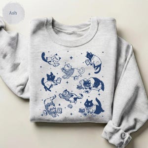 May include: An ash gray crewneck sweatshirt featuring a dark blue graphic print of multiple cats in various poses, including some wearing lab coats and glasses, surrounded by small star shapes, books, and scientific documents.