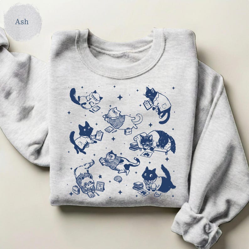 May include: An ash gray crewneck sweatshirt featuring a dark blue graphic print of multiple cats in various poses, including some wearing lab coats and glasses, surrounded by small star shapes, books, and scientific documents.