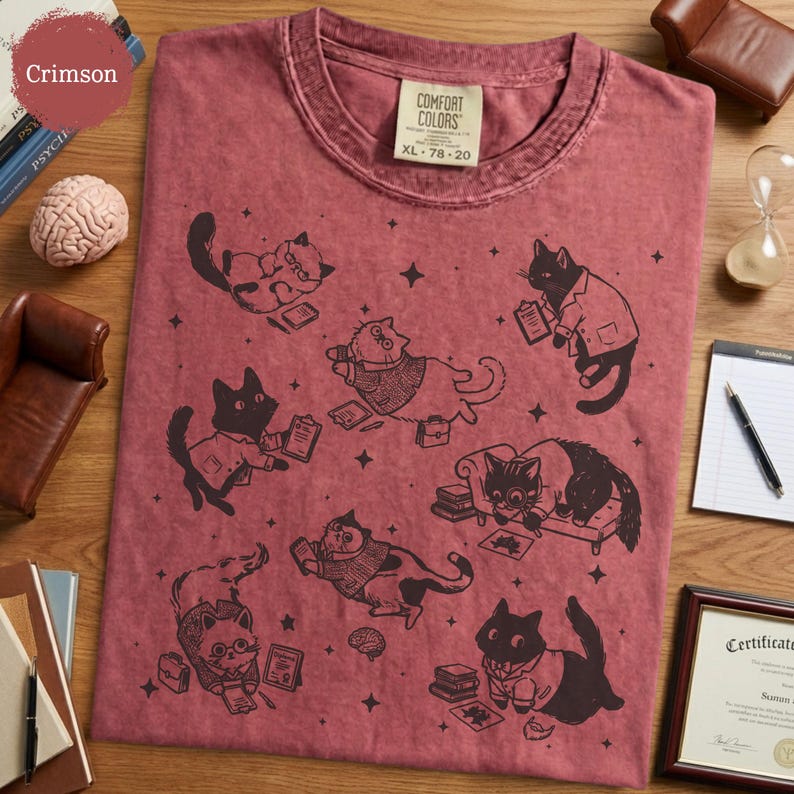 May include: A crimson t-shirt featuring a black graphic print of various cats in professional attire, including cats with glasses, lab coats, and briefcases, interspersed with small stars and books. The shirt has a Comfort Colors brand label.