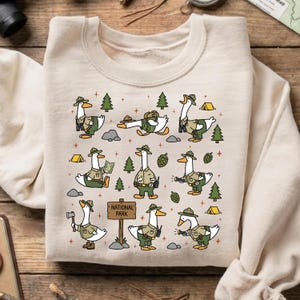Op de afbeelding: Beige sweatshirt met een herhaald patroon van cartoon ganzen verkleed als parkwachters. De ganzen worden in verschillende poses afgebeeld, met bomen, tenten en de tekst "National Park".