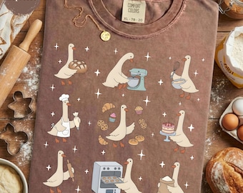 Op de afbeelding: Een bruin t-shirt met korte mouwen en een grafische print van witte ganzen die aan het bakken zijn. De ganzen zijn afgebeeld terwijl ze mixen, kneden en taarten versieren, omringd door kleine witte sterren en gebak.