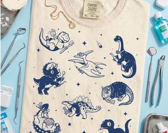 Könnte beinhalten: Ein elfenbeinfarbenes T-Shirt mit einem dunkelblauen Grafikdruck verschiedener Dinosaurier, die mit Zahnarztinstrumenten interagieren. Das Shirt hat ein Comfort Colors-Etikett am Kragen mit der Größenangabe XL. Das Design zeigt Sterne und Dinosaurier.