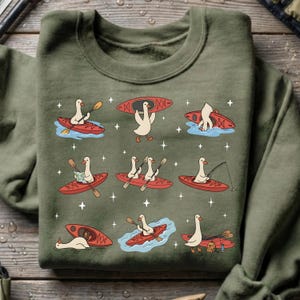 May include: An olive green sweatshirt featuring a whimsical design of cartoon geese kayaking. The geese are depicted in red kayaks, engaged in various activities like paddling, fishing, and carrying their kayaks. The design includes water and star accents.
