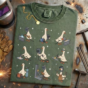 May include: Green t-shirt featuring a whimsical design of cartoon geese welding. The geese are depicted in various welding poses with tools and equipment. The shirt has a vintage look and feel.