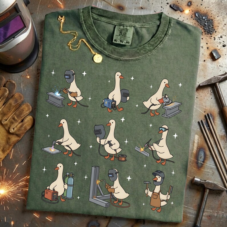 May include: Green t-shirt featuring a whimsical design of cartoon geese welding. The geese are depicted in various welding poses with tools and equipment. The shirt has a vintage look and feel.