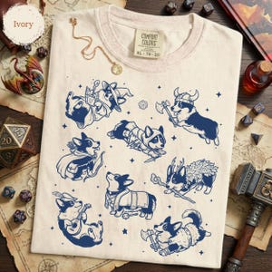 May include: An ivory t-shirt featuring a dark blue graphic print of various corgi dogs dressed as fantasy characters, including a wizard, a viking, and a bard. The shirt has a Comfort Colors brand label at the neck with size XL 78-20 printed on it.