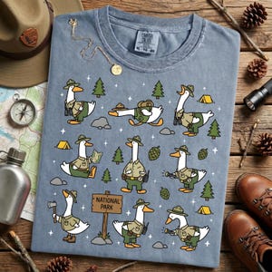 Op de afbeelding: Blauw Comfort Colors T-shirt met een speels ontwerp van cartoon ganzen gekleed als parkwachters. De ganzen worden getoond in verschillende buitenscenario's, waaronder wandelen, kaarten lezen en kamperen. Het shirt heeft een "National Park" bord.