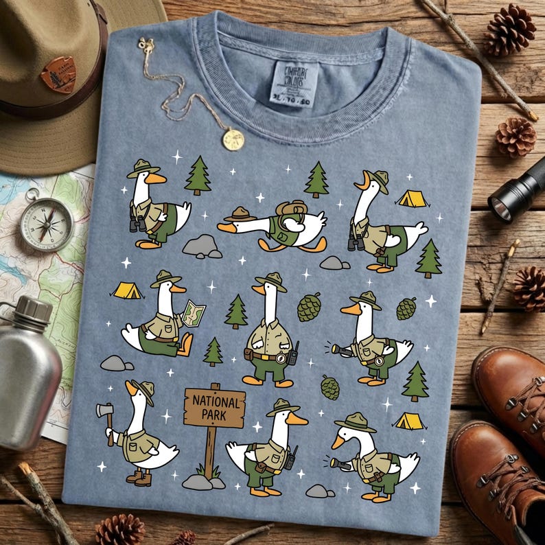 Op de afbeelding: Blauw Comfort Colors T-shirt met een speels ontwerp van cartoon ganzen gekleed als parkwachters. De ganzen worden getoond in verschillende buitenscenario's, waaronder wandelen, kaarten lezen en kamperen. Het shirt heeft een "National Park" bord.